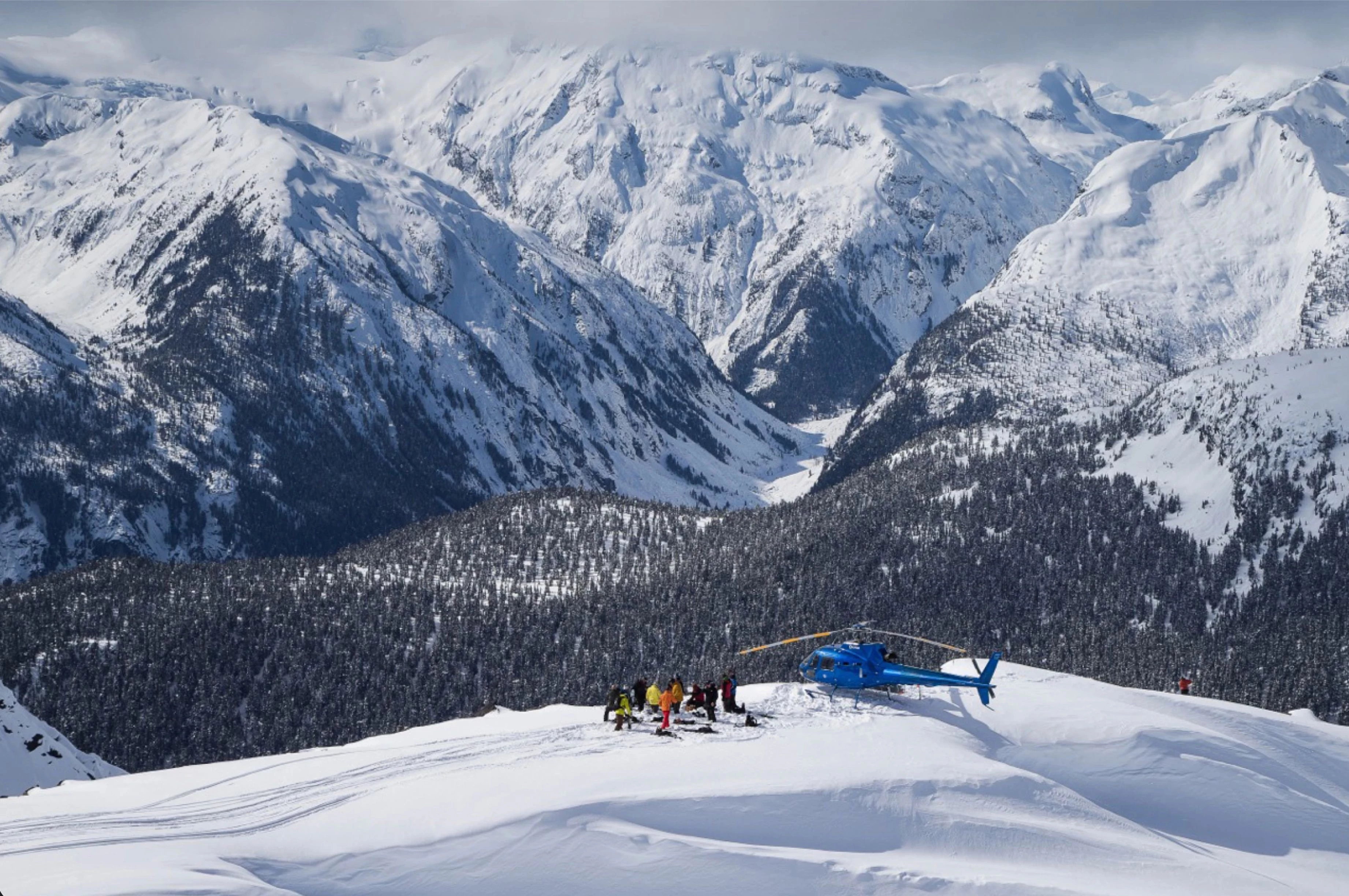 Skiers and a helicopter on a snowy ridge overlooking a forested valley in the Skeena Range.