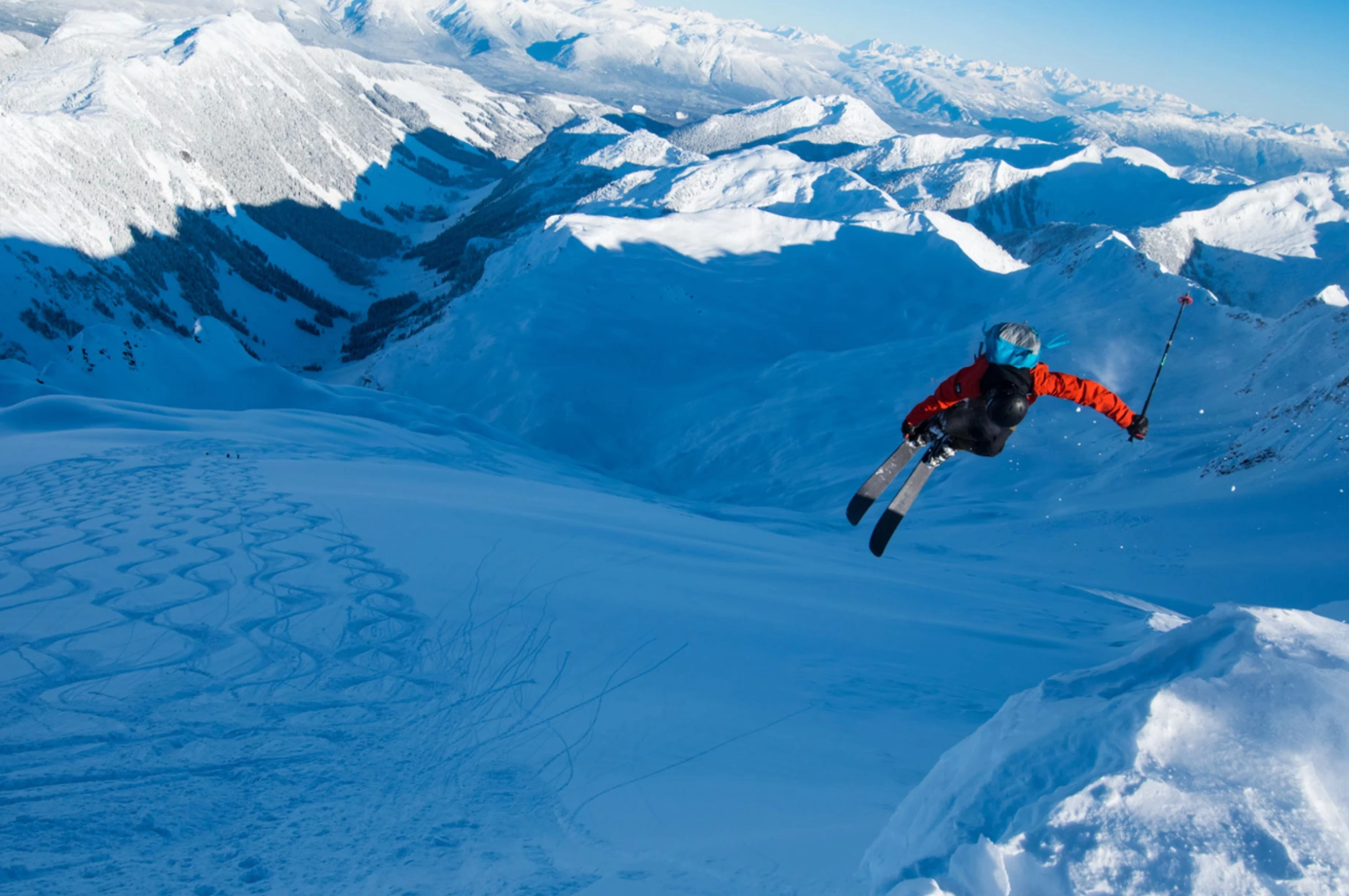 A skier in mid-air above a wide, snow-covered alpine bowl surrounded by mountain peaks.