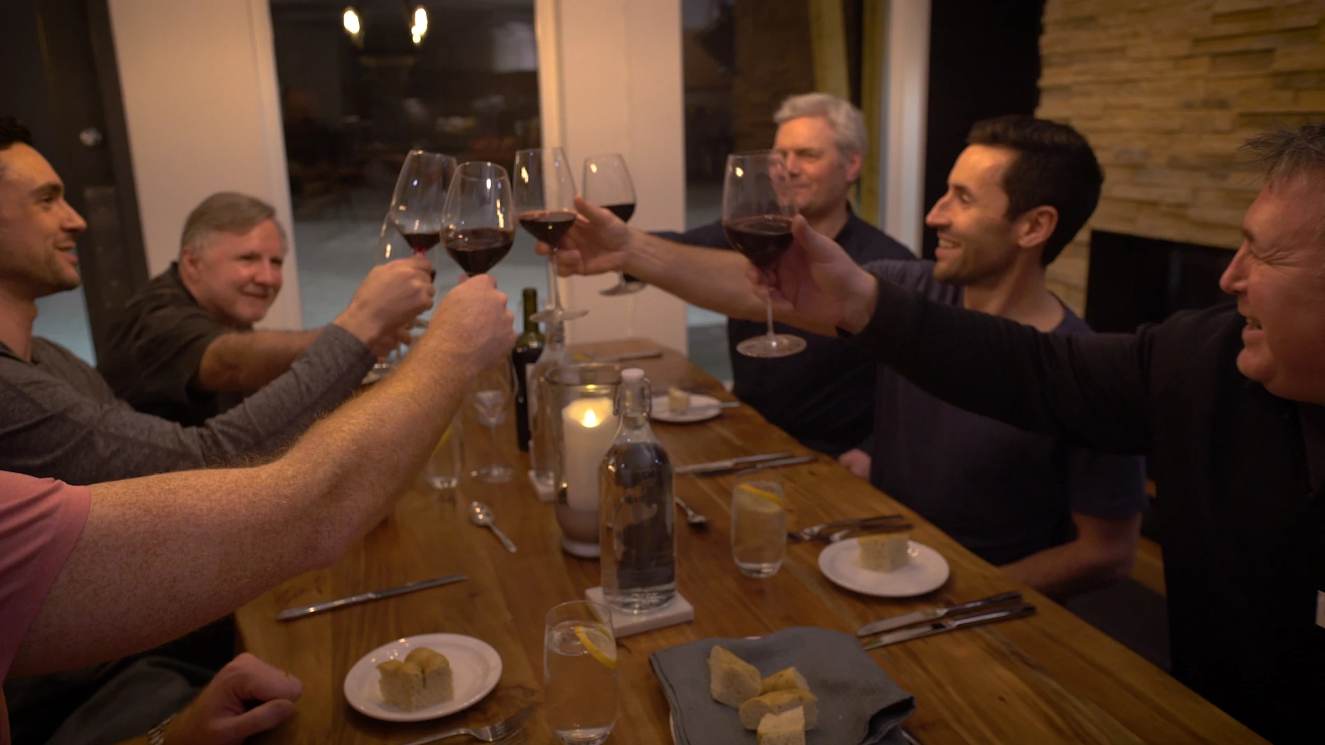 Six men clink red wine glasses in a toast around a long wood dinner table in a lodge dining room, with a stone fireplace, candlelight and bread plates set out.