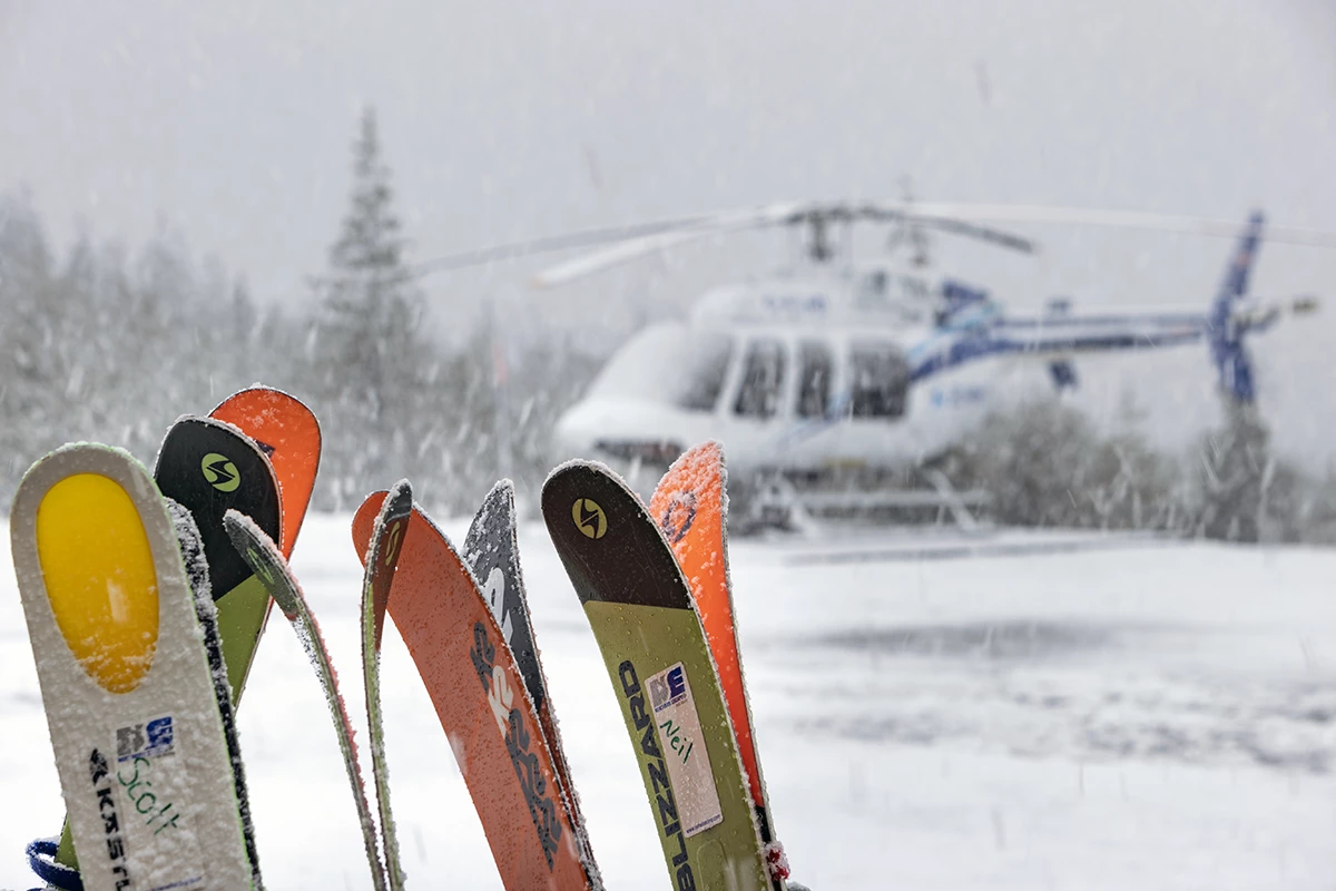 Skis lined up in the snow as a helicopter lands in the background during a snowfall in the Skeena Mountains.